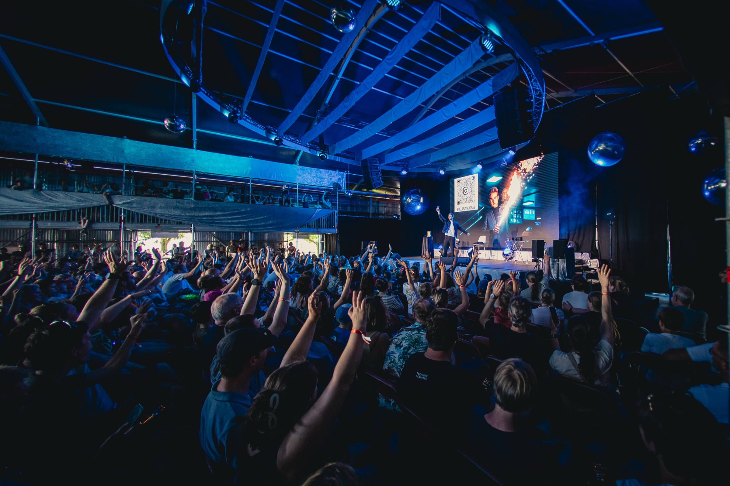 A large audience with hands raised during a stage performance under blue lighting